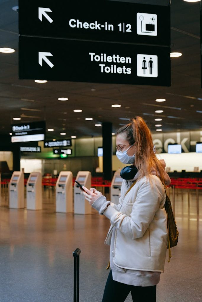 Woman wearing a mask, texting in airport terminal with signage in background.